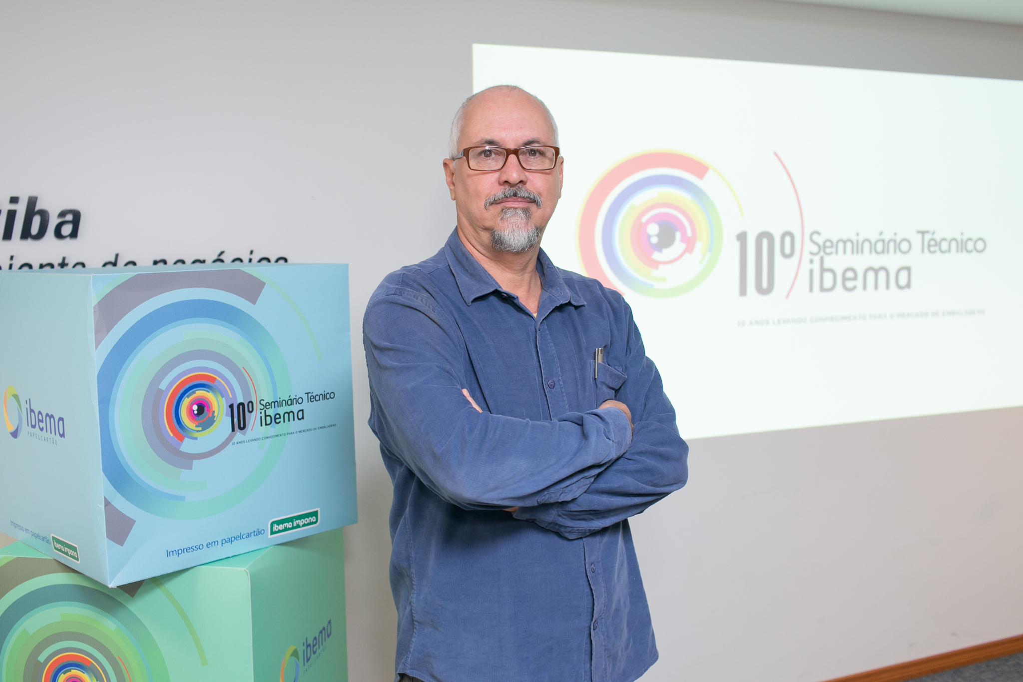 Na imagem, o designer Fábio Mestriner está com braços cruzados em frente à uma projeção de slides onde se lê "10º Seminário Técnico IBEMA", empresa onde realiza consultoria.