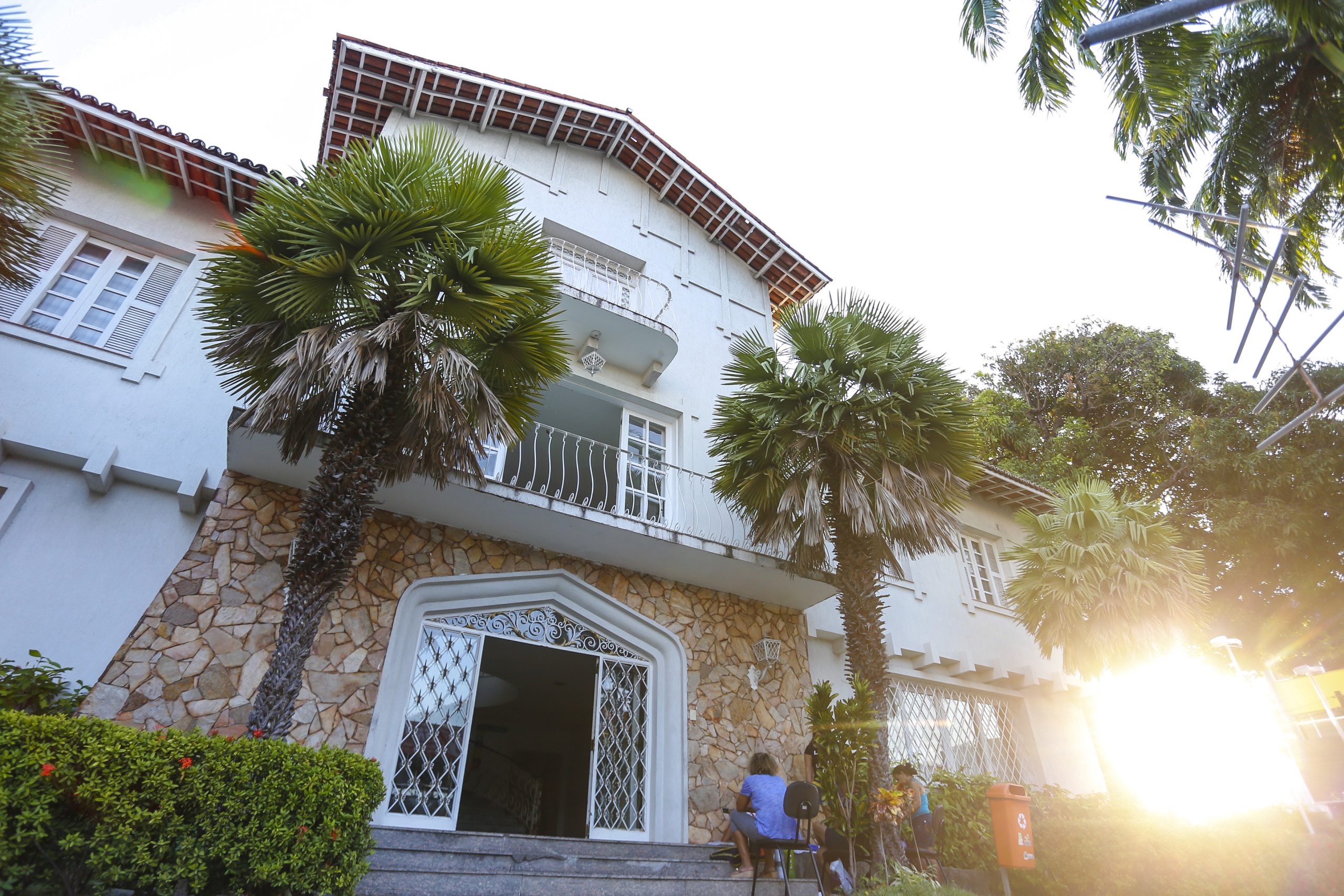 Fachada da Vila das Artes vista de baixo para cima. Além da casa, podem-se ver coqueiros e muitas plantas e, à direita, a luz do sol e duas pessoas sentadas.