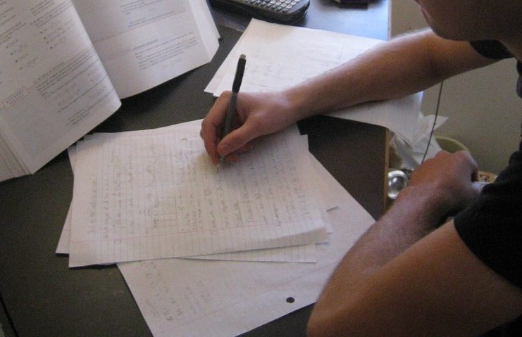 Mão de um rapaz fazendo anotações em folhas de papel brancas; há um livro em pé e aberto na mesa, que é também material necessário para cursinho comunitário