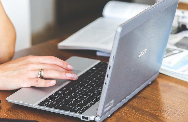 Uma mão de uma mulher com três anéis no dedo anelar teclando em um computador cinza com o teclado preto, ao fundo tem livros abertos em cima da mesa de madeira marrom que também apoia o computador