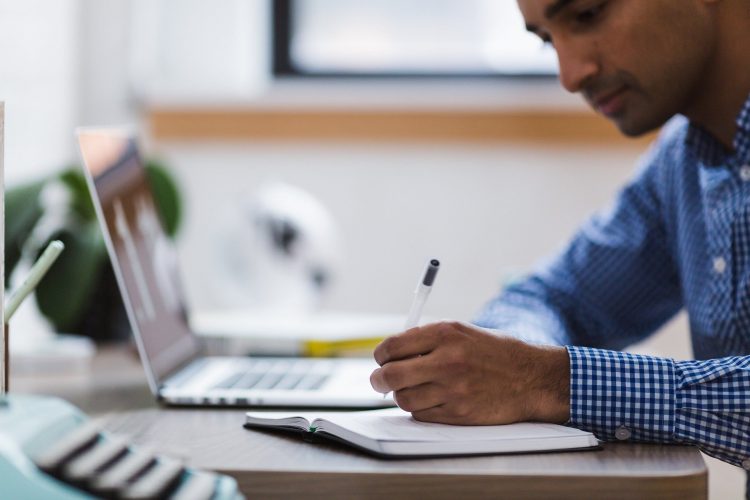 Homem de blusa azul trabalha sobre uma mesa e anota em um caderno. Há um notebook ao seu lado. Ele parece praticar home office