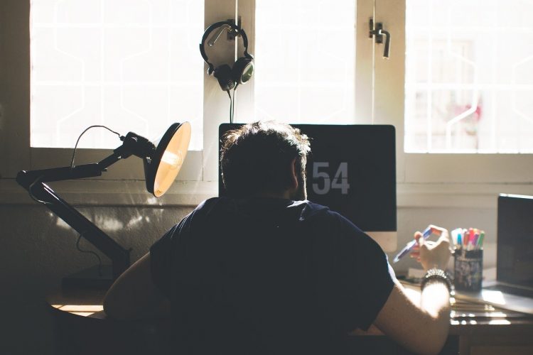 Homem sentado em frente ao computador, com bloco de notas e caneta e na mão; manter a produtividade durante o home office é desafio para profissionais