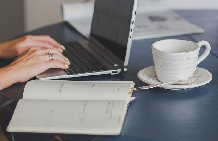 Mãos brancas digitando em um laptop, com xícara de café e caderno sobre a mesa; futuro do mercado corporativo será discutido em webinar online