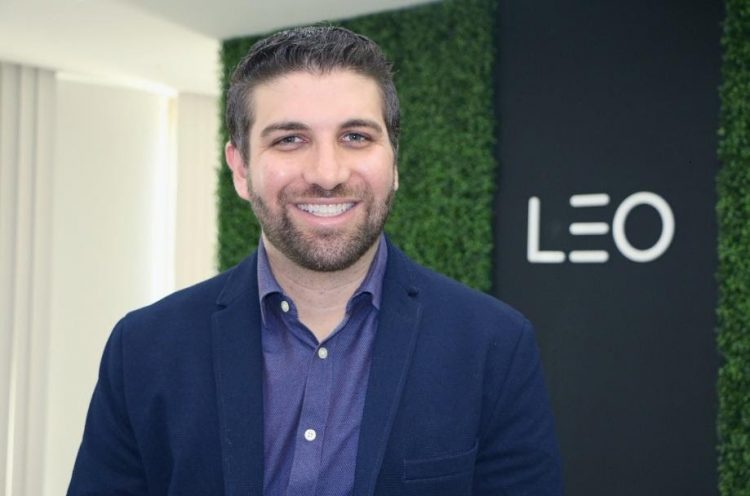 Na foto, o CEO da LEO Learning, Richard Vasconcelos, sorri para a câmera. Ele usa uma camisa social azul e um blazer também azul em um tom mais escuro. Usa barba, é branco e está em frente a um painel onde se lê "LEO"
