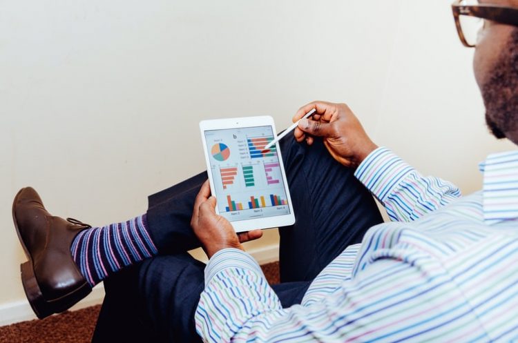 Senac Comércio - curso explorará tendências para empresários. Na foto, há um homem de roupa social com um tablet na mão. No tablet, há vários gráficos expostos, aparentemente relacionados à finanças. A meia do homem, listrada, é aparente