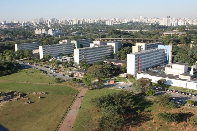 A foto é uma imagem aérea do campus da USP na cidade de São Paulo. Há quatro prédios grandes e brancos, separados por pátios e árvores