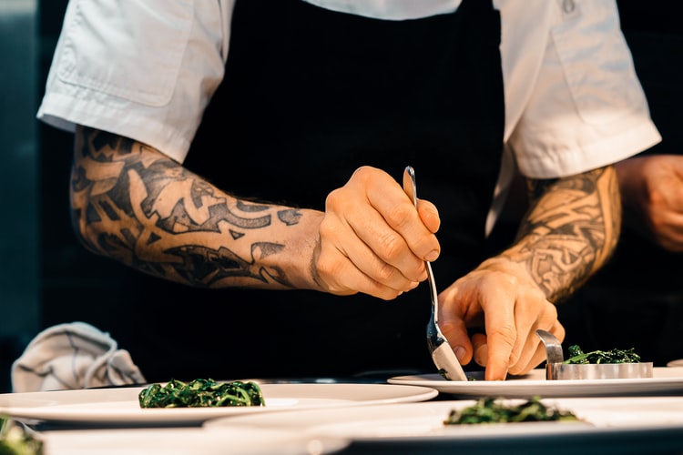 Gastronomia: a foto mostra um chef preparando a apresentação de pratos. Os dois braços do homem, que é branco, são tatuados. O rosto não está visível, apenas uma camisa branca e um avental preto.