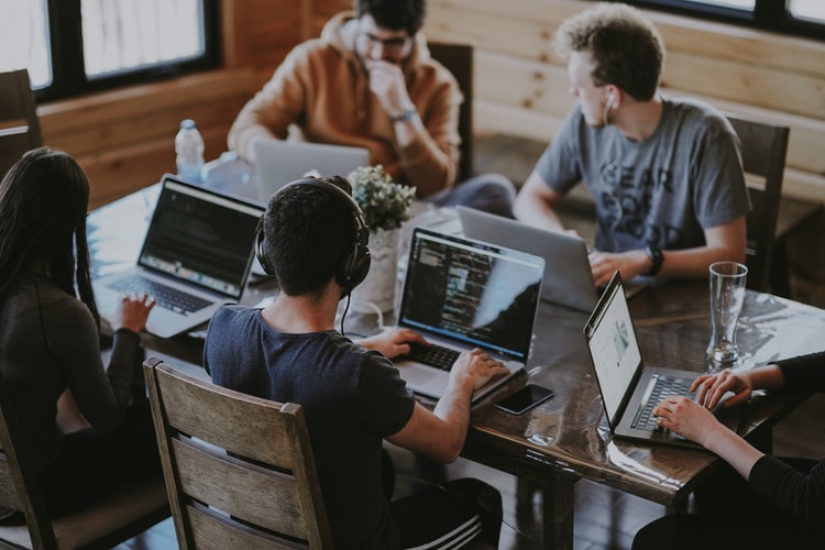 Aceleração: na imagem, um grupo de cinco jovens discute ideias e acessa a internet em notebooks. São quatro homens e uma mulher. O ambiente é claro e eles se reúnem em uma mesa de madeira.