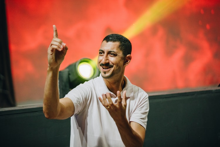 Na imagem, um homem jovem de camisa branca e cabelo e bigode pretos interpreta sinais. Atrás, há um holofote e luzes vermelhas, que indicam um show com acessibilidade.