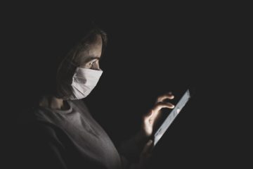 Covid-19: uma mulher de máscara mexe em um tablet no escuro. Ela está de perfil e usa a mão esquerda para manipular o equipamento.