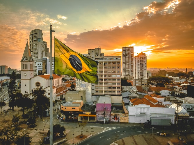 Curso sobre pós-pandemia no Brasil: na imagem, há uma paisagem urbana na luz do pôr-do-sol. Em primeiro plano, há uma bandeira do Brasil tremulando.