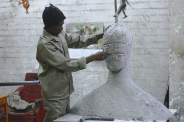 Capacitação para agentes culturais: na imagem, há um homem negro com um macacão bege e uma touca preta. Ele está esculpindo uma estátua que possui formato de busto, mas ainda não possui face.