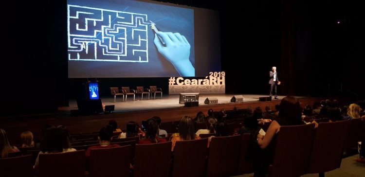 CearáRH: Na imagem, uma palestra do evento de 2019. O público está em um auditório escuro e, no palco, há um telão e uma escultura com o nome "#CearáRH" em branco.