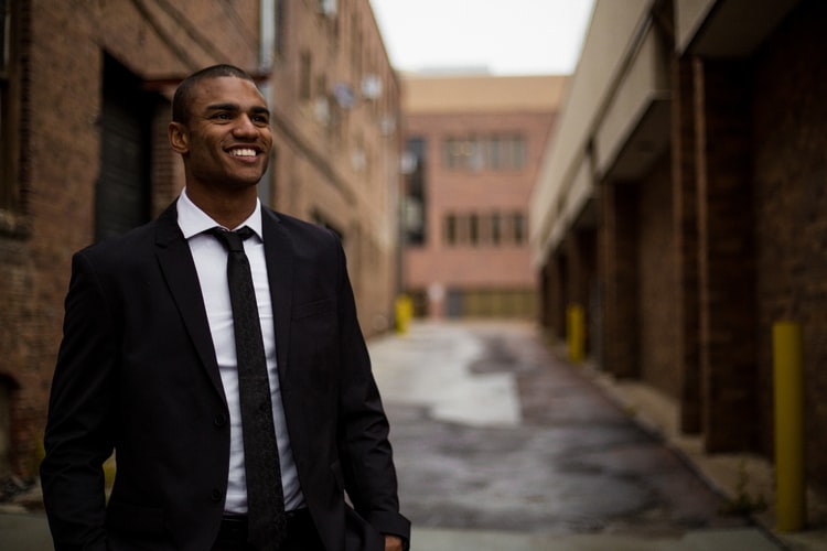 Lideranças negras: na imagem, um jovem negro sorri. Ele usa terno e gravata pretas e uma camisa social de cor branca e posa em frente à uma rua vazia.