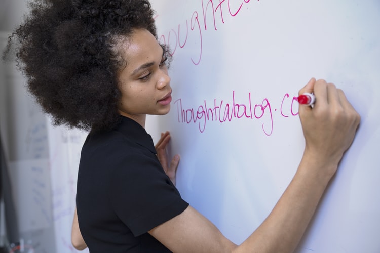 Semana Pedagógica: a imagem mostra uma mulher negra e jovem escrevendo em um quadro branco. Ela usa camisa de manga curta preta, cabelo solto e crespo e escreve com uma caneta de cor vermelha.