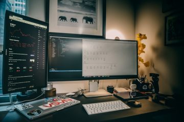 Vagas home office: imagem mostra um pequeno escritório improvisado em casa, com diversos computadores com gráficos em cima de uma mesa. Atrás da mesa há um quadro preto e branco e, por trás dos computadores, uma luminária.