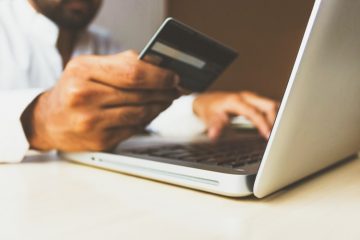 E-commerce: na imagem, um par de mãos segura um cartão de crédito em frente a um computador. Não é possível ver o rosto do comprador.